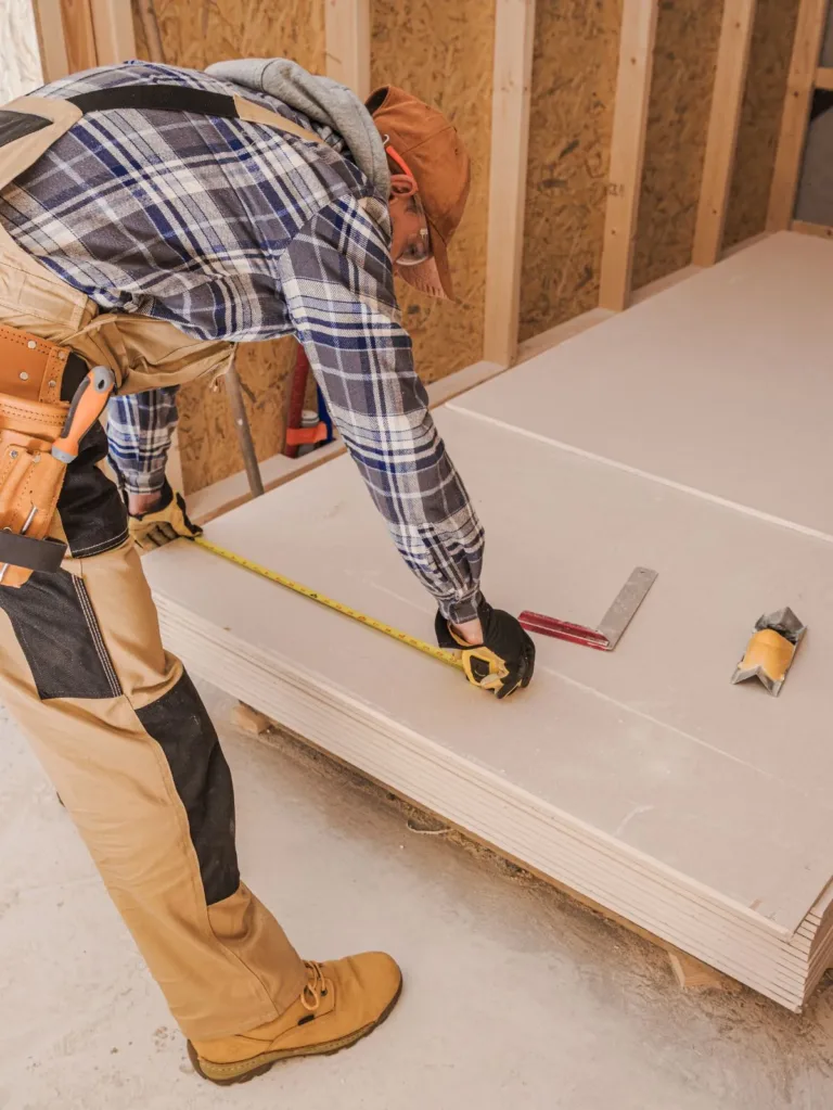 A construction worker in a flannel shirt and work boots measures wooden panels, illustrating Bayside Builders Group's expertise in precise home renovation projects across the Bay Area.